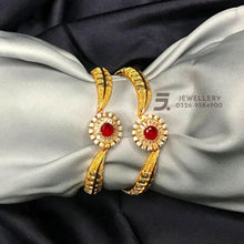 Gold bangles with red stones on a gray fabric background, brand 'Jewellery' visible.